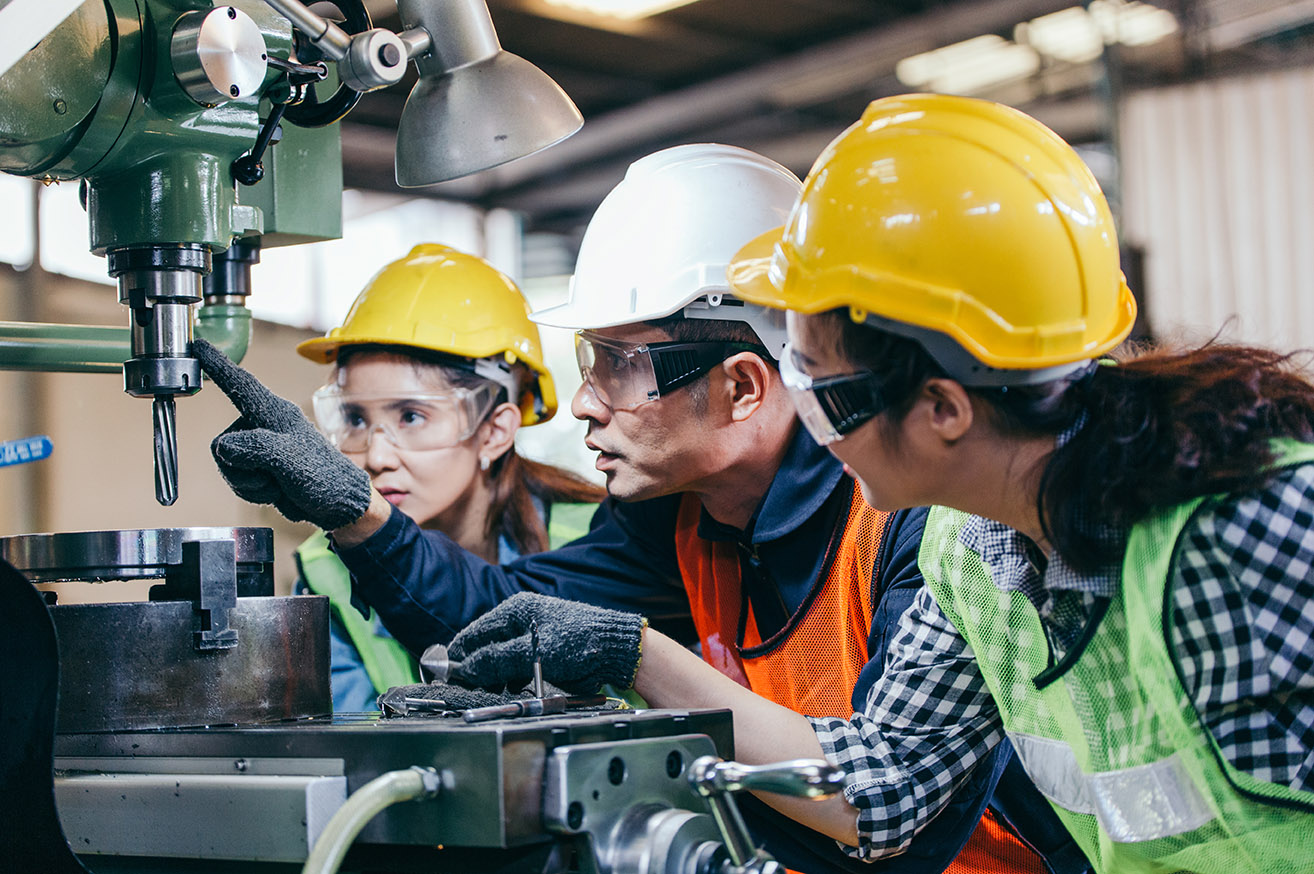 Asian male foreman manager showing case study of factory machine to two engineer trainee young woman in protective uniform.